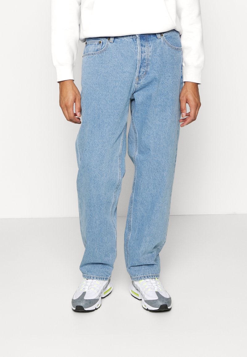 Lyseblå denimjeans med løs passform. Frontlukk med knapp og glidelås, fem lommer og synlige sømdetaljer. Kombinert med hvite og grå sneakers.