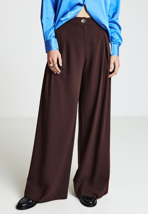Pantalon marron à jambes larges avec une texture lisse, doté d'un pli à l'avant et d'un bouton métallique distinctif à la taille. Chaussures noires visibles.