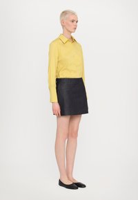 Camicia gialla a maniche lunghe con bottoni abbinata a una mini gonna in denim scuro. L'outfit presenta un colletto classico e dettagli discreti nelle tasche.