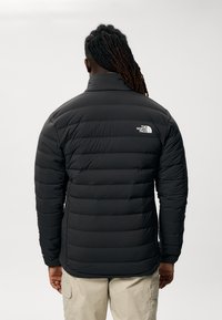 Casaco de puffer preto com design acolchoado horizontal, gola alta e pequeno logo branco na parte superior das costas. Textura suave e acolchoada.