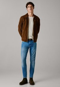 Brązowa skórzana kurtka na jasnym t-shircie, w połączeniu z jasnoniebieskimi dżinsami i ciemnobrownymi botkami za kostkę. Prosty design, luźny krój, minimalne detale.