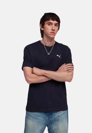 Uomo con capelli corti e scuri che indossa una t-shirt Puma blu navy e jeans azzurri, braccia incrociate, su uno sfondo chiaro uniforme.