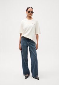 Mujer vestida con una camiseta blanca de gran tamaño con el texto "Park Avenue", jeans de pierna ancha azules, cinturón negro, zapatos de punta con tachuelas y gafas de sol negras.