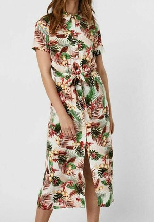 Femme portant une robe boutonnée à manches courtes avec une taille nouée, présentant un imprimé floral tropical aux tons verts, marron et rouges.