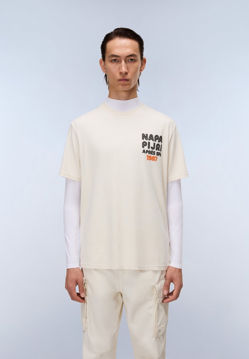 Krèmefarvet bomulds t-shirt med korte ærmer, med sort og orange tekst på venstre side, lagdelt over en hvid langærmet skjorte.