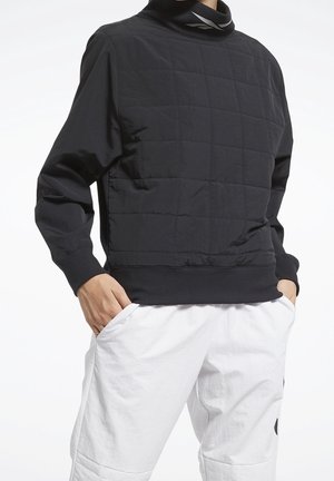 Pullover nero oversize con texture trapuntata, collo alto e polsini elasticizzati, abbinato a pantaloni bianchi con tasca laterale.