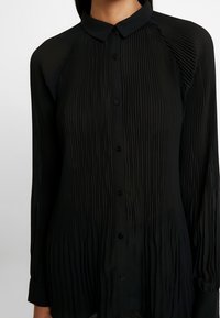 Maglietta nera a maniche lunghe con un design plissettato e testurizzato. Presenta un colletto classico e chiusura con bottoni, con una leggera lucentezza e un orlo curvo.