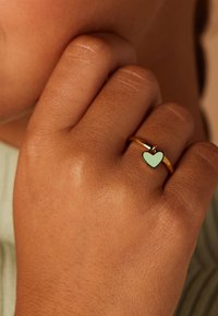 Gouden ring met een mintgroene hartcharme. De hand heeft een gladde, getextureerde huid en houdt de ring dicht bij het gezicht.