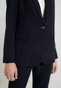 Veston de costume bleu marine avec un motif à pois rouges et bleus. Doté d'un revers cranté, d'une fermeture à un bouton, et d'une poche avant. Le tissu présente une texture apparente.