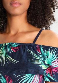 Bluzka z odkrytymi ramionami wykonana z granatowej tkaniny z kolorowymi tropikalnymi liśćmi w zielonym, różowym i czerwonym kolorze. Regulowane cienkie ramiączka.