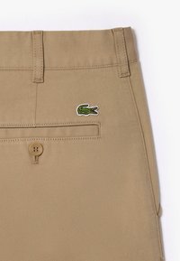 Calças de algodão bege com um acabamento texturizado, apresentando um bolso traseiro e um pequeno logótipo de crocodilo verde bordado no lado direito.