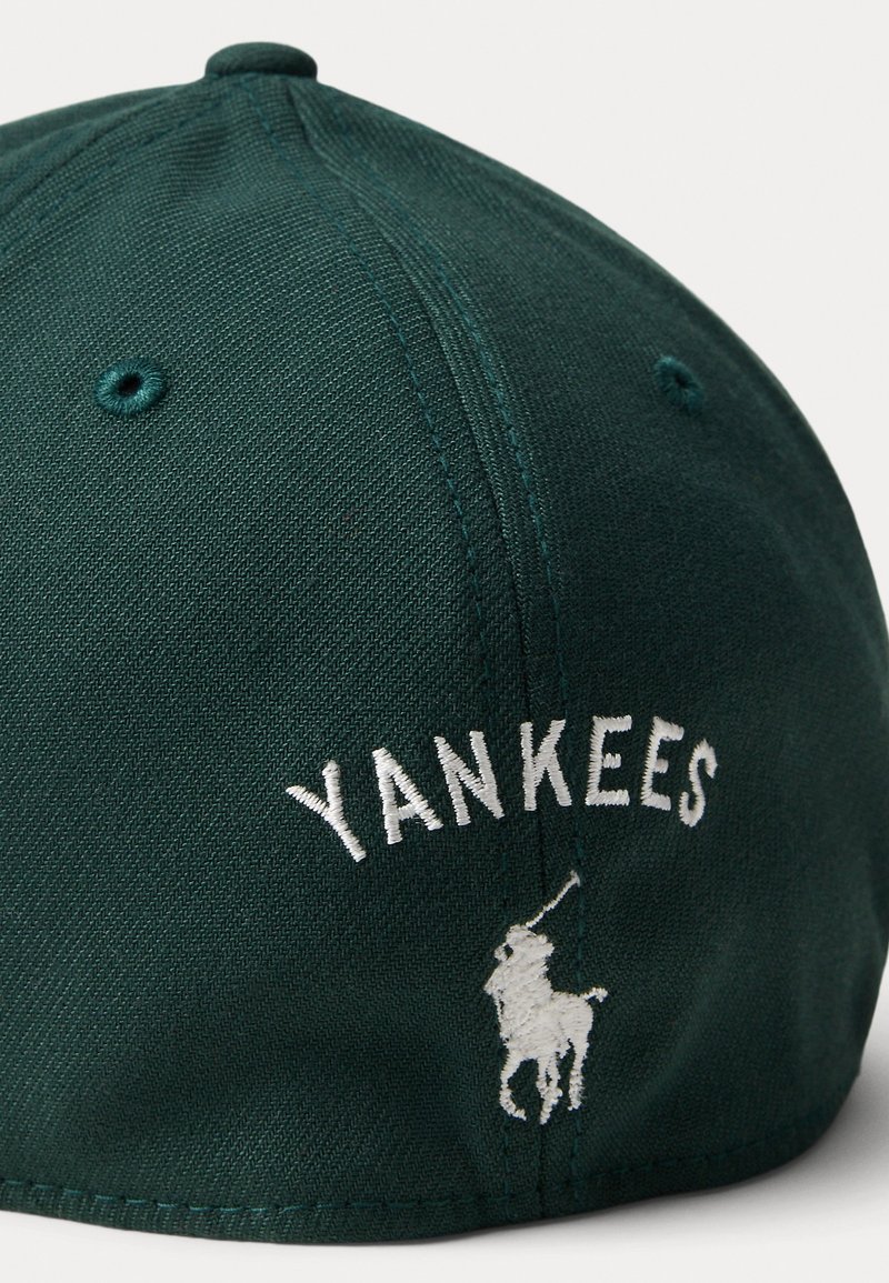 Mörkgrön basebollkeps med vit broderad text "YANKEES" och en liten vit polo-spelare logotyp på sidan. Strukturerad tyg, böjd skärm.