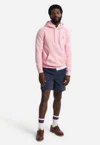 Felpa rosa chiaro con tasca frontale, abbinata a pantaloni corti blu navy. Scarpe marroni e calze bianche con strisce rosse e blu. Uomo in piedi con le mani rilassate.