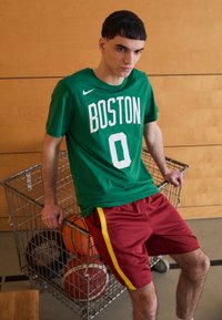 Grøn kortærmet Nike T-shirt med "BOSTON" og "0" i hvid. Røde shorts med gule sider, stående ved en trådkurv fyldt med basketballer.