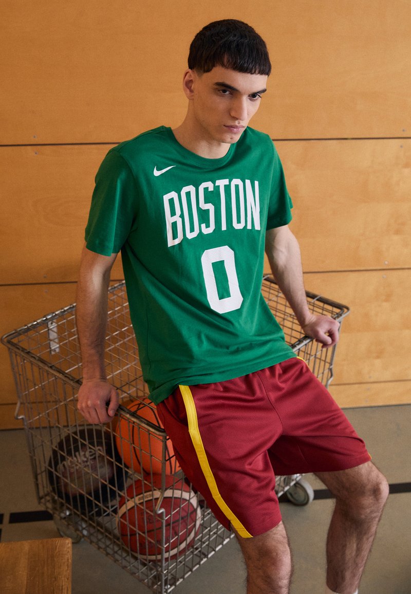 Grøn kortærmet Nike T-shirt med "BOSTON" og "0" i hvid. Røde shorts med gule sider, stående ved en trådkurv fyldt med basketballer.