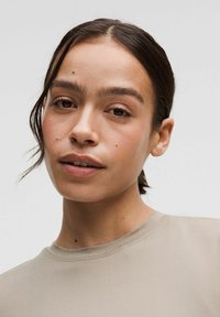 Beige t-shirt with a round neckline; smooth fabric; subtle stitching details; model has light skin and dark hair, shown against a plain background.