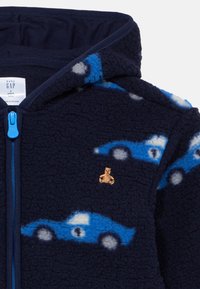 Φούτερ με κουκούλα σε ναυτικό μπλε χρώμα με σχέδια μπλε αυτοκινήτων, χρυσό έμβλημα αρκούδας και μπλε φερμουάρ. Μαλακή υφή και άνετος σχεδιασμός κατάλληλος για νήπια.