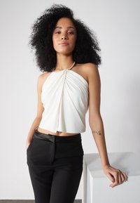 Balts halter top ar savērpamu dizainu un metāliskiem kakla rotājumiem, apvienots ar pieguļošām melnām biksēm, uz vienkārša fona.