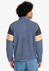 Polarowy pulower w kolorze niebieskim, z poziomymi paskami w kolorach granatowym i beżowym. Zamek błyskawiczny do połowy wysokości oraz teksturowana tkanina. Aktywny krój z elastycznymi mankietami.