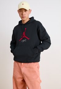 Hanorac negru cu glugă și logo roșu Jordan, buzunar tip cangur și șnur. Capac galben pal cu logo roșu. Pantaloni roz deschis.