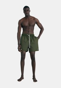 Bañador verde oscuro con cintura elástica, cordón blanco y un logo en el dobladillo. El modelo está descalzo y de pie.