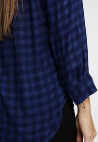 Långärmad blå rutigt skjorta bärs över svarta byxor, med synlig handledstatuering på personens högra arm.