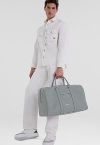 Sac de voyage gris avec un motif texturé, deux poignées supérieures, et une poche zippée à l'avant, associé à une chemise oversize blanche et un pantalon.