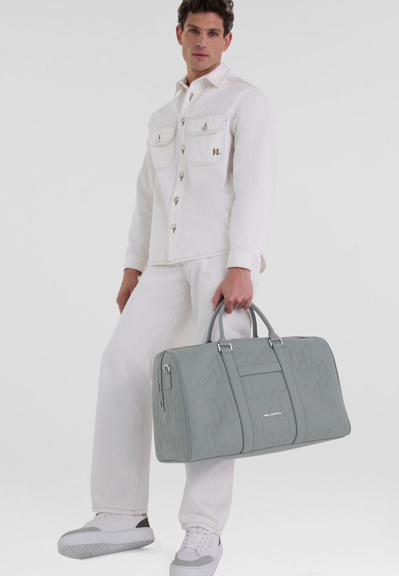 Sac de voyage gris avec un motif texturé, deux poignées supérieures, et une poche zippée à l'avant, associé à une chemise oversize blanche et un pantalon.