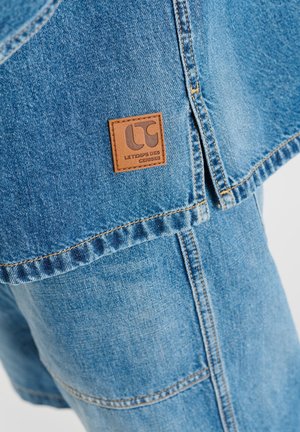 Blaue Denimjacke mit braunem Lederetikett mit der Aufschrift „Le Temps des Cerises“ über passenden Jeanshosen.