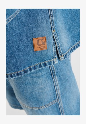 Blaue Denimjacke mit braunem Lederetikett mit der Aufschrift „Le Temps des Cerises“ über passenden Jeanshosen.