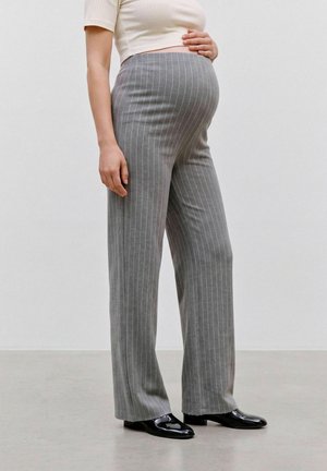 Persona incinta che indossa pantaloni premaman grigi a righe sottili, top crema a coste e scarpe nere lucide, in piedi davanti a uno sfondo semplice.