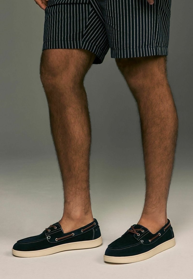Jambes portant un short à rayures noires et blanches et des chaussures décontractées noires à enfiler avec des semelles blanches sur un fond neutre.