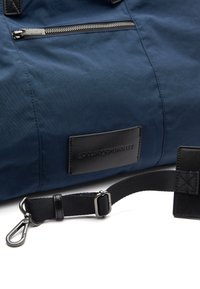 Borsa in tessuto blu navy con tasca con zip, superficie textured, dettagli in pelle nera, tracolla regolabile e un'etichetta con logo cucita sul lato.