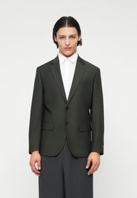 Dunkelgrüne maßgeschneiderte Blazer mit zwei vorderen Knöpfen, Reverskragen und seitlichen Taschen, kombiniert mit einem weißen Hemd und grauen Hosen.