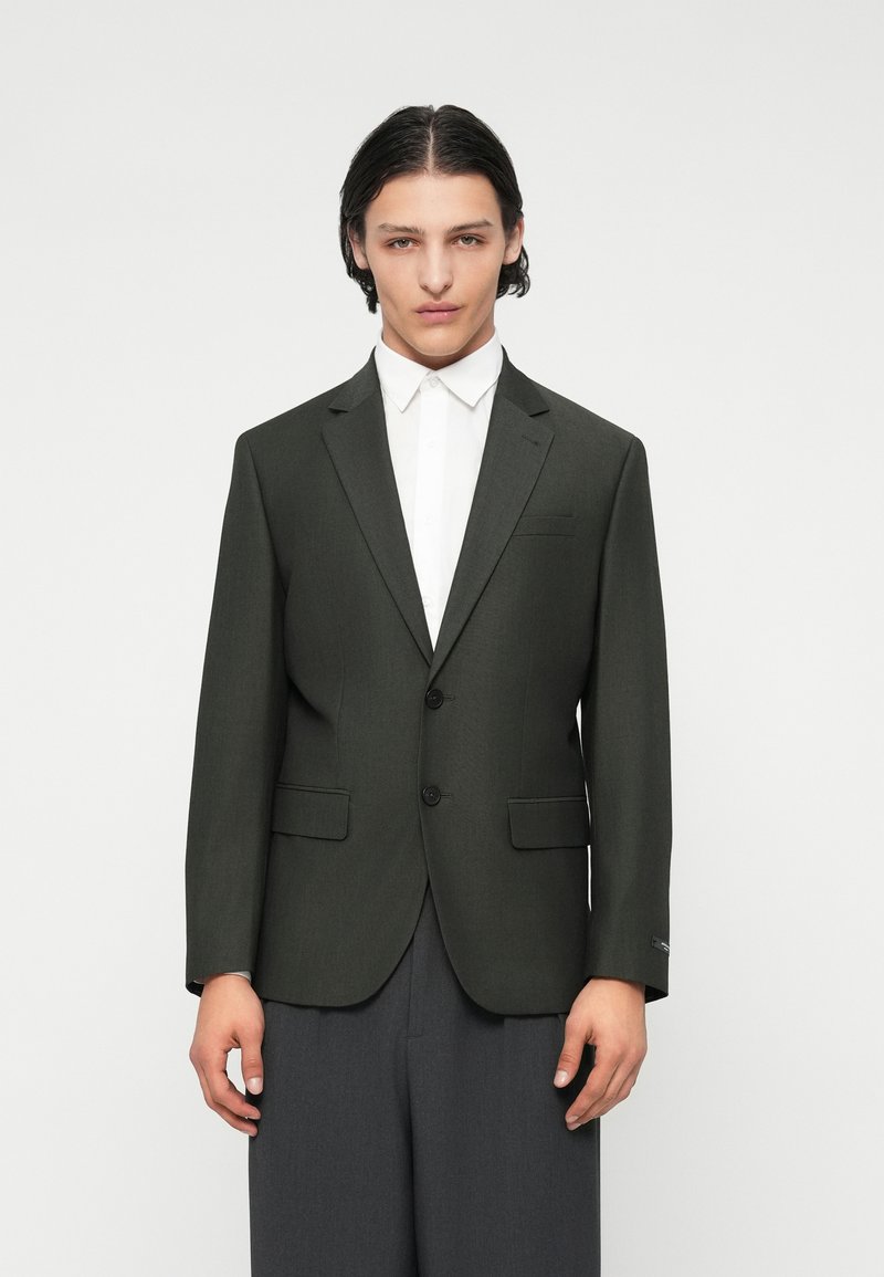 Dunkelgrüne maßgeschneiderte Blazer mit zwei vorderen Knöpfen, Reverskragen und seitlichen Taschen, kombiniert mit einem weißen Hemd und grauen Hosen.