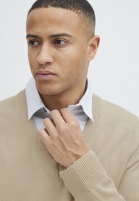 Beige V-Ausschnitt-Pullover mit gerippten Bündchen über einem hellblau-weiß gestreiften Hemd mit Kragen, das einen Knopf am Hals aufweist.
