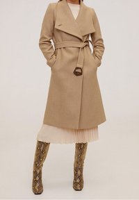 Manteau à double boutonnage en mélange de laine beige avec ceinture et col classique, associé à une robe plissée claire et des bottes hautes à imprimé serpent.