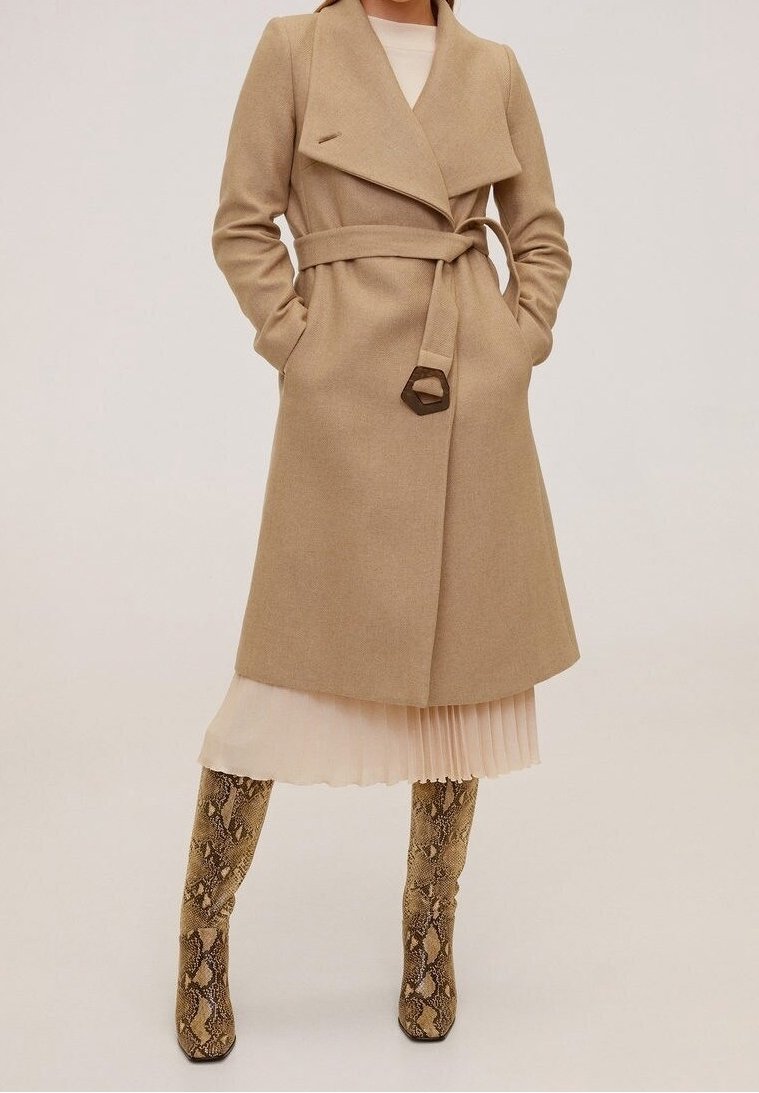 Manteau à double boutonnage en mélange de laine beige avec ceinture et col classique, associé à une robe plissée claire et des bottes hautes à imprimé serpent.