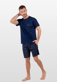 Marinblå bomulls t-shirt med en dekorativ ficka, tillsammans med marinblå shorts med ett blommönster i röd och blå, barfota.