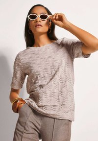 Texturiertes Taupe-T-Shirt mit Wellenmuster, Rundhalsausschnitt und kurzen Ärmeln. Kombiniert mit hellgrauen Hosen und übergroßen weißen Sonnenbrillen.
