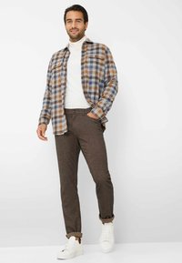 Geruite flanellen shirt in blauw, bruin en wit; witte coltrui; bruine op maat gemaakte broek; witte sneakers; model staat met handen in de zakken.