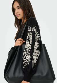 Grote zwarte leren boodschappentas met een soepele textuur, versierd met ingewikkelde witte borduursels op de mouw van een zwart sweater die door een persoon wordt vastgehouden.