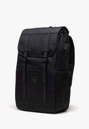 Sac à dos noir au design structuré, en matériau textile, avec des bretelles ajustables, une poche avant et un rabat replié sécurisé par des boucles.