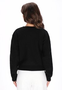 Fekete kötött pulóver laza szabással, esővállel és bordás mandzsettával. A textúra puhanak tűnik, enyhe fénycsillogással. A modellen fehér alsó öltözet van.