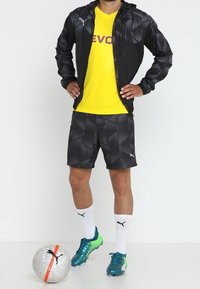 Träningskläder bestående av en svart mönstrad jacka, gul skjorta med texten "EVOC", svarta shorts och turkosa fotbollsskor. Silverfärgad fotboll med röd rand.