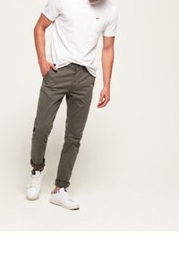 T-shirt bianca in cotone con taschino sul petto, abbinata a pantaloni verde oliva e sneakers bianche. I pantaloni presentano orli arrotolati e una vestibilità slim.