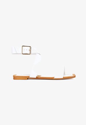 Sandalias planas blancas con dos cintas anchas y cierre de hebilla. La suela es color marrón claro, con un acabado suave y elementos de diseño mínimos.