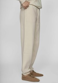 Pantalones de chándal beige, de corte holgado, con cintura y puños elásticos. Material suave, bolsillos laterales y textura lisa. Combinados con pantuflas marrones y peludas.
