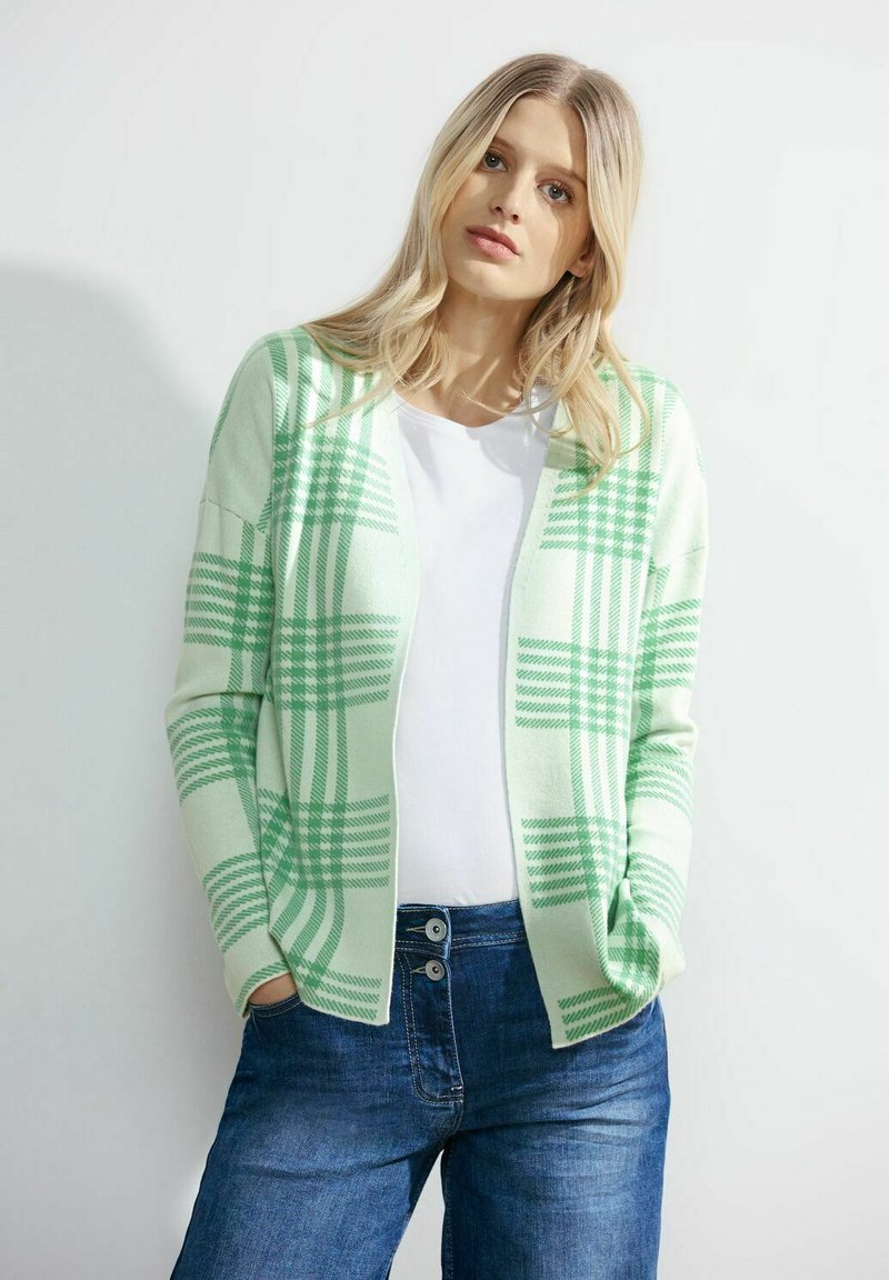 Světle zelený kostkovaný cardigan s jemnou texturou, otevřený přední design, dlouhé rukávy, nošený přes bílou košili a doplněný modrými džíny.