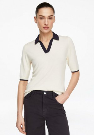 Femme portant un polo crème à manches courtes avec col et poignets noirs, associé à un pantalon noir taille haute, main dans la poche.